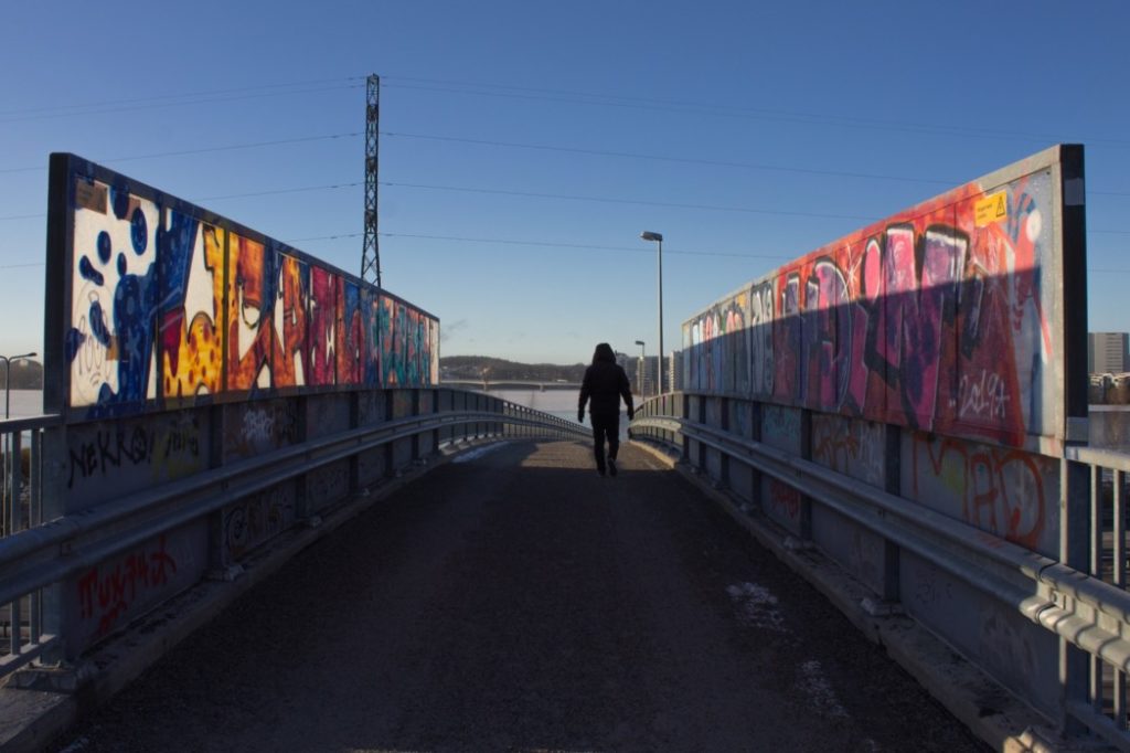 ihminen kävelee sillalla, silta on maalattu värikkäillä graffiteilla, taivas on sininen, talvi