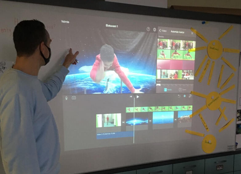 Mies seisoo luokan edessä ja osoittaa taulua, jossa näkyy videotyökaluohjelma